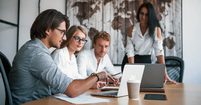 Four professionals in business attire collaborate around a laptop in a modern office, reviewing contract data—symbolizing teamwork and digital contract management.