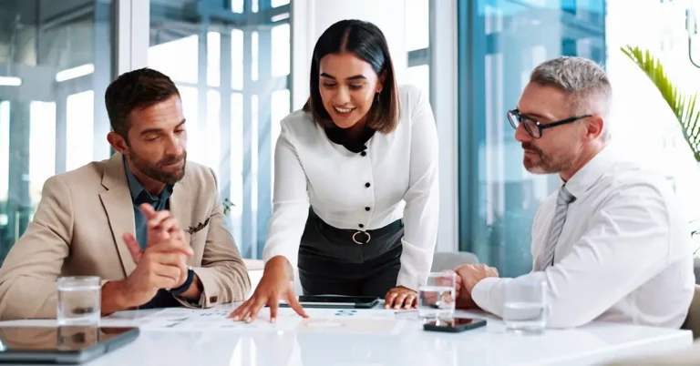 “Three professionals in a modern office reviewing documents together. A woman in business attire leads the discussion, pointing to a paper on the table while two men listen attentively. Represents collaboration and strategic planning.