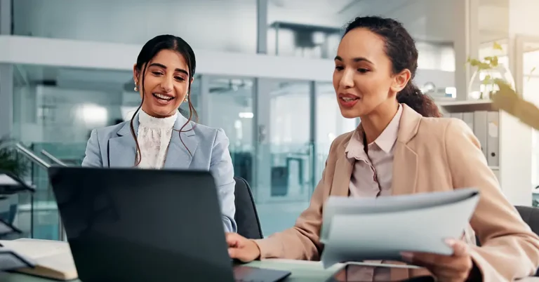 Two professional women in business attire review documents together at a laptop in a modern office, representing collaborative AI document review solutions.