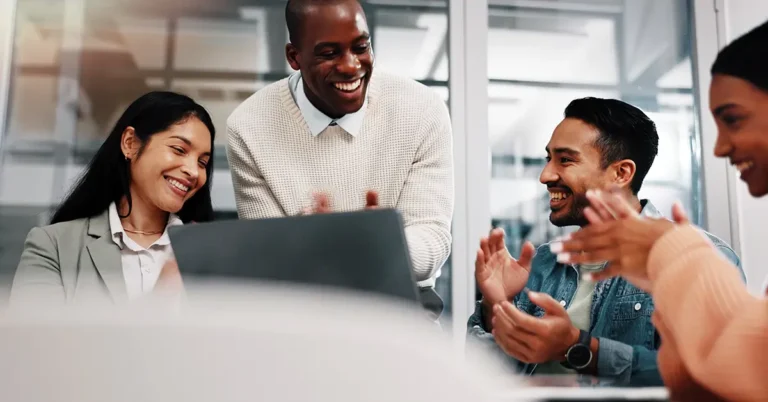 Group of professionals smiling and clapping around a laptop in a modern office, symbolizing collaboration and success with Leah Tariff Agent.