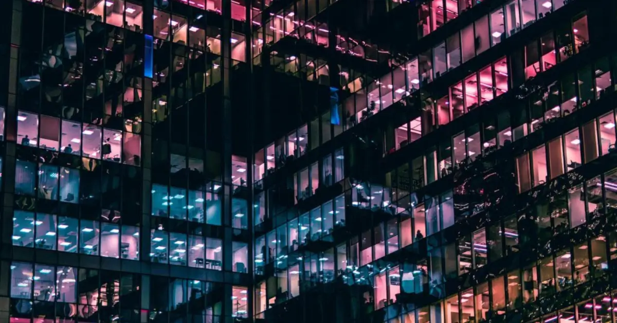Nighttime view of a modern office building with glass windows lit in pink, blue, and purple tones, revealing silhouettes of workers inside.
