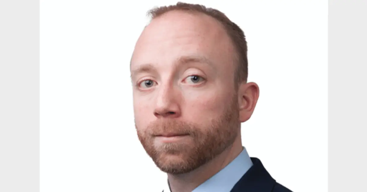 Professional headshot of a man with short light brown hair and beard, wearing a navy suit and light blue shirt against a white background.