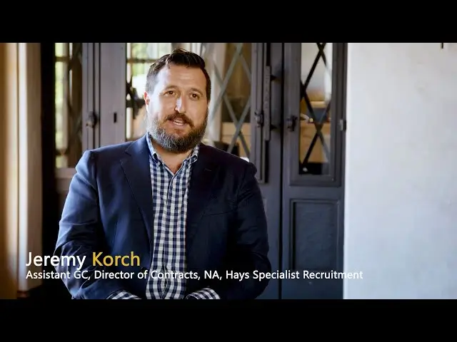 Jeremy Korch, Assistant GC and Director of Contracts at Hays Specialist Recruitment, speaks during an interview wearing a navy blazer and checkered shirt.