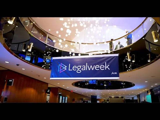 Large blue Legalweek banner by ALM hangs in a modern conference venue with elegant lighting and people visible on the upper floor balcony.