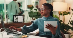 Professional woman in glasses sits at modern office desk using a laptop and holding a tablet, symbolizing leadership and digital transformation.