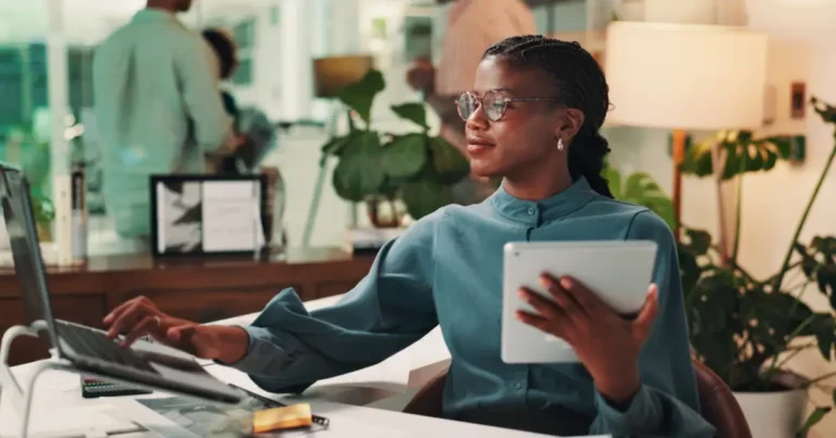 Professional woman in glasses sits at modern office desk using a laptop and holding a tablet, symbolizing leadership and digital transformation.