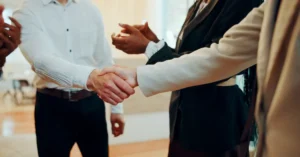 Close-up of a professional handshake between two businesspeople in formal attire, with colleagues applauding in the background, symbolizing leadership growth and partnership.