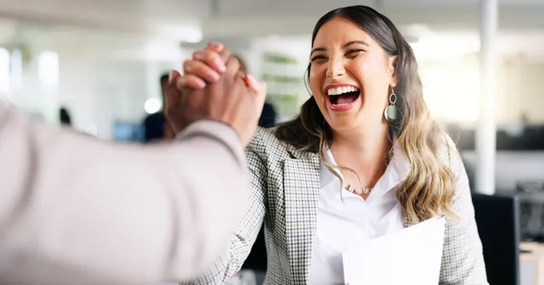 Smiling businesswoman in a blazer and earrings celebrating a win with a handshake in a bright office, symbolizing leadership and customer success.